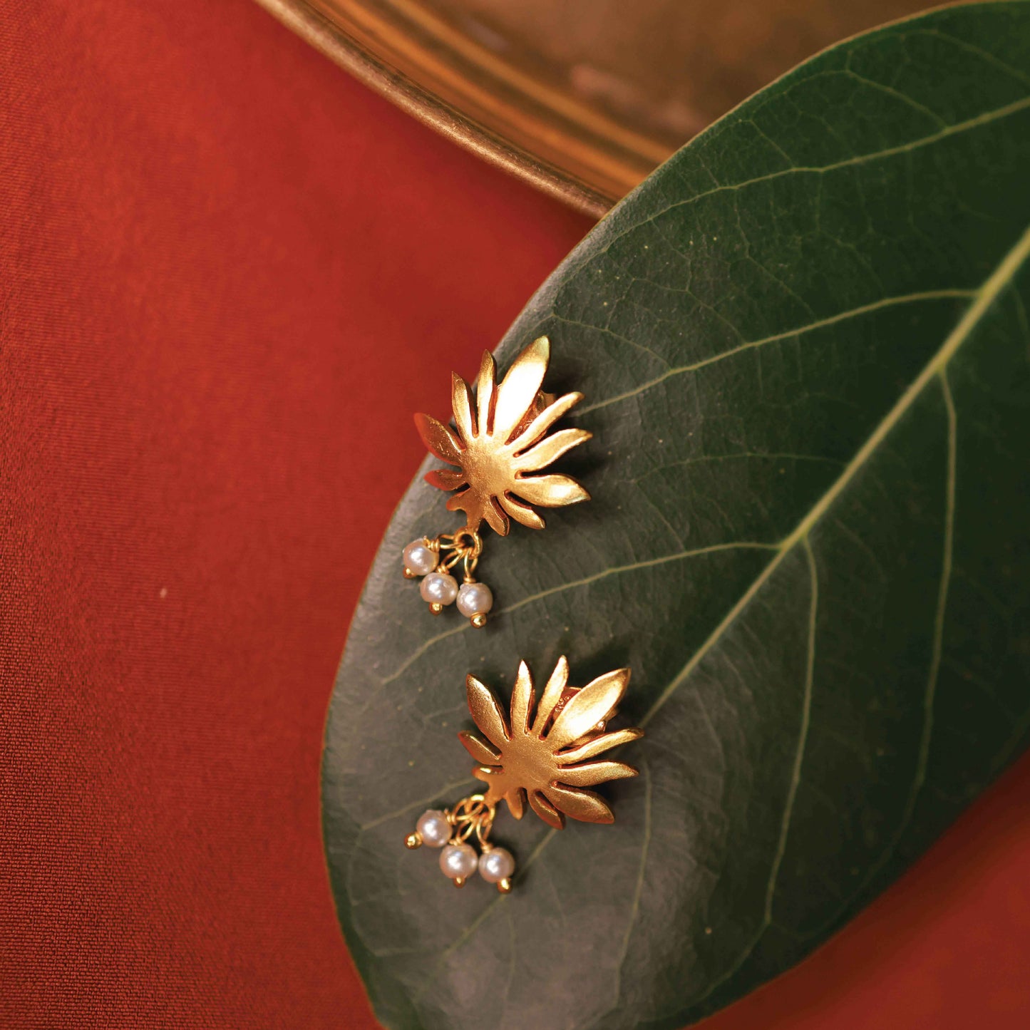 Gold floral earrings with pearl accents on a green leaf against a red fabric background