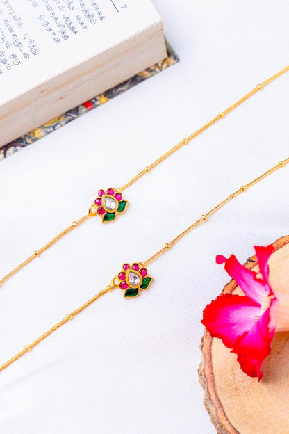 Gold anklets with floral designs on a white surface with a book and pink flower petal.