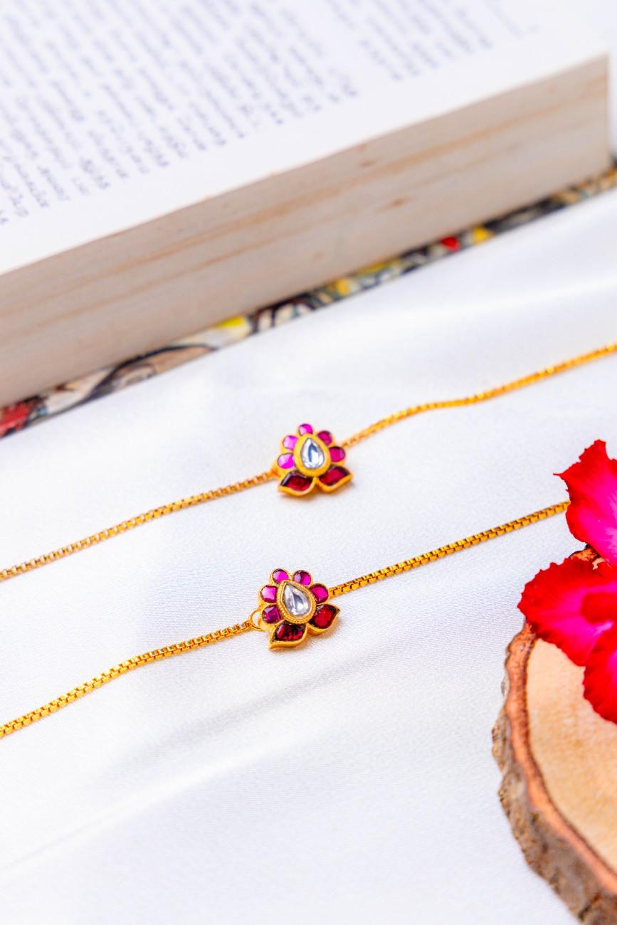 Two gold plated anklet with red and gold pendants on a white surface with a book and wooden slice in the background.