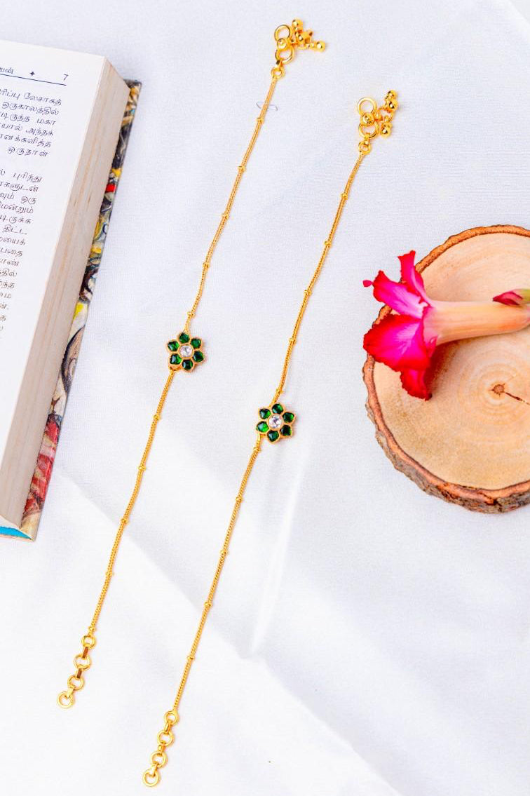Gold plated anklet with green gemstones on a white surface with a book and pink flower.