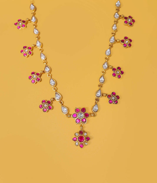 Gold-plated necklace with pink gemstone floral motifs and teardrop-shaped clear stones, displayed on a solid yellow background.