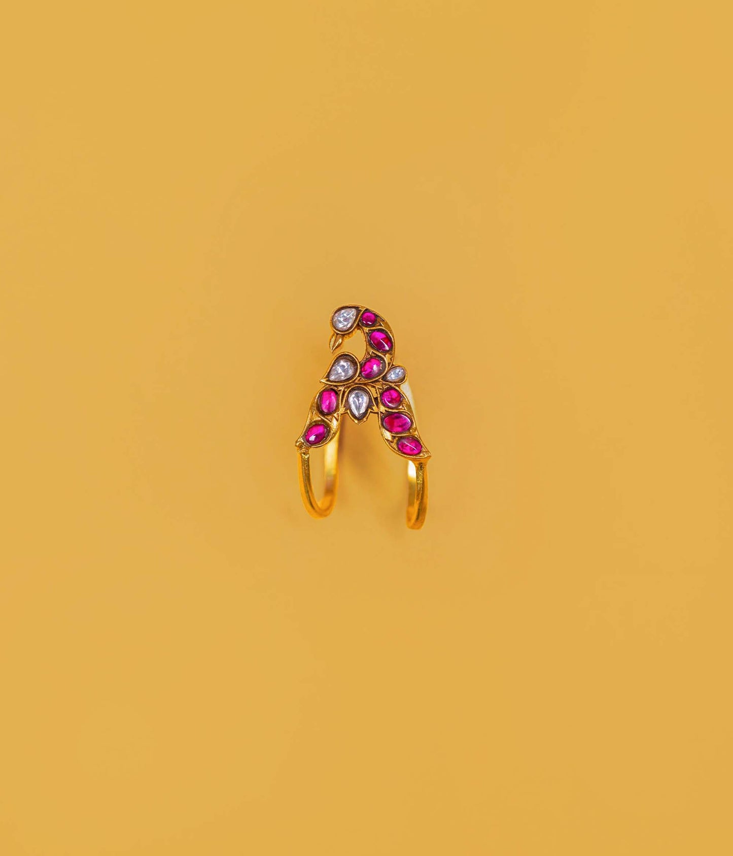 A gold-plated split ring featuring a decorative top with pink teardrop-shaped stones, a white teardrop stone, and a small round clear stone, set against a solid mustard yellow background.