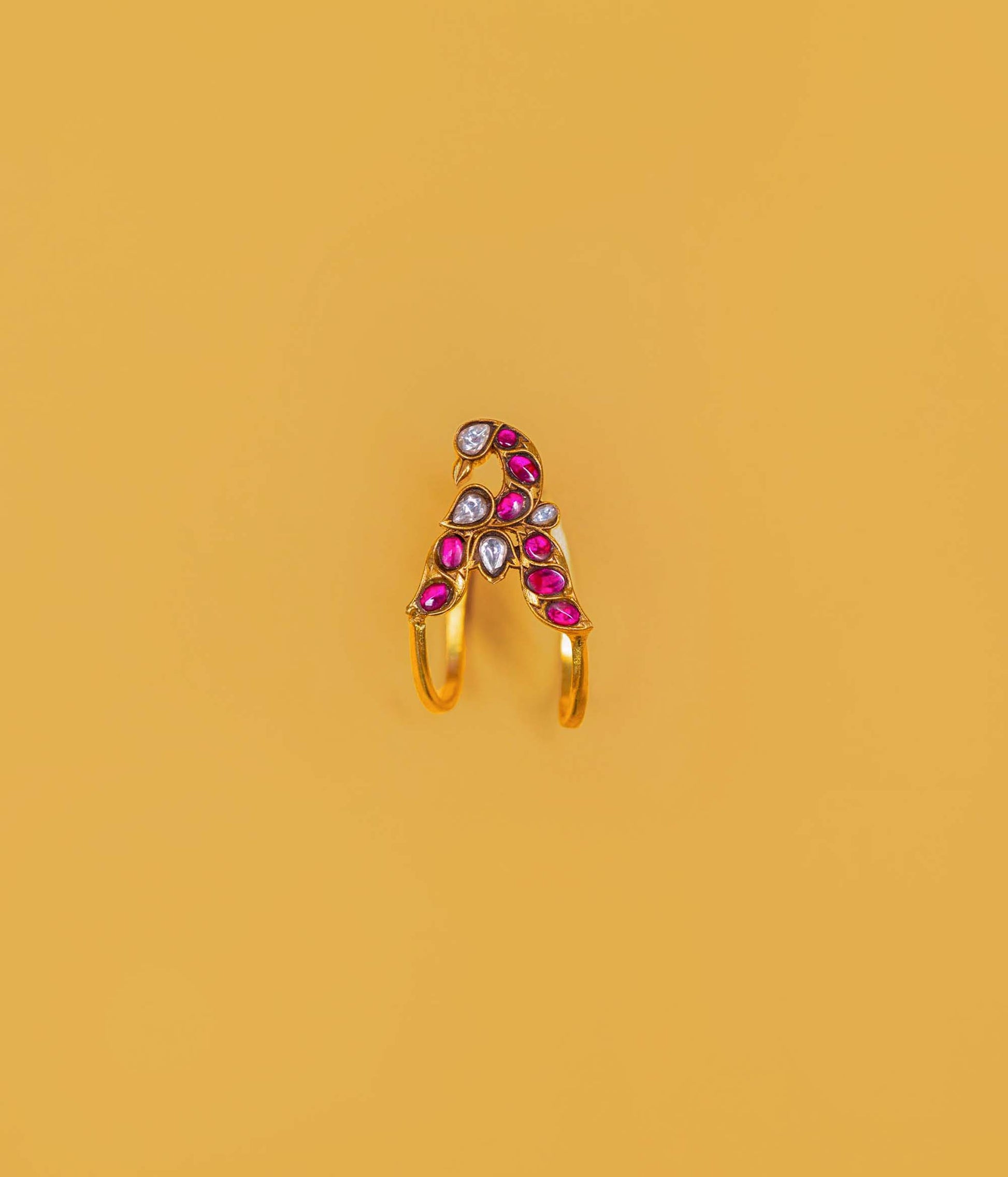 A gold-plated split ring featuring a decorative top with pink teardrop-shaped stones, a white teardrop stone, and a small round clear stone, set against a solid mustard yellow background.