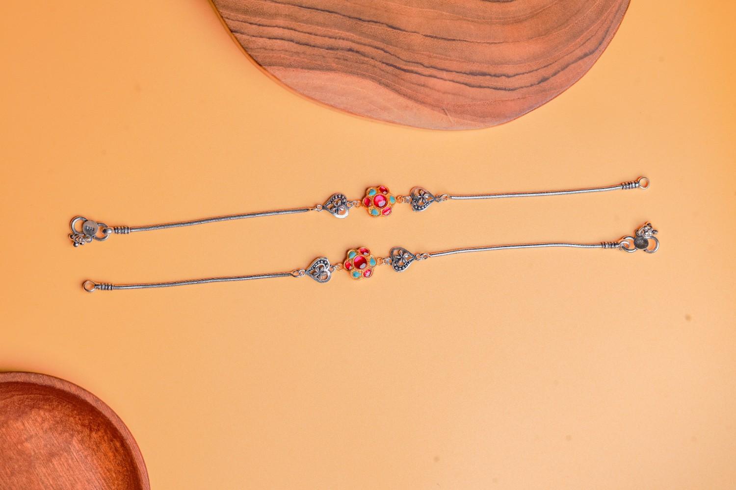 Dual-tone silver anklets with floral and heart motifs, red and green Kundan stones by Sil’alankaraa.