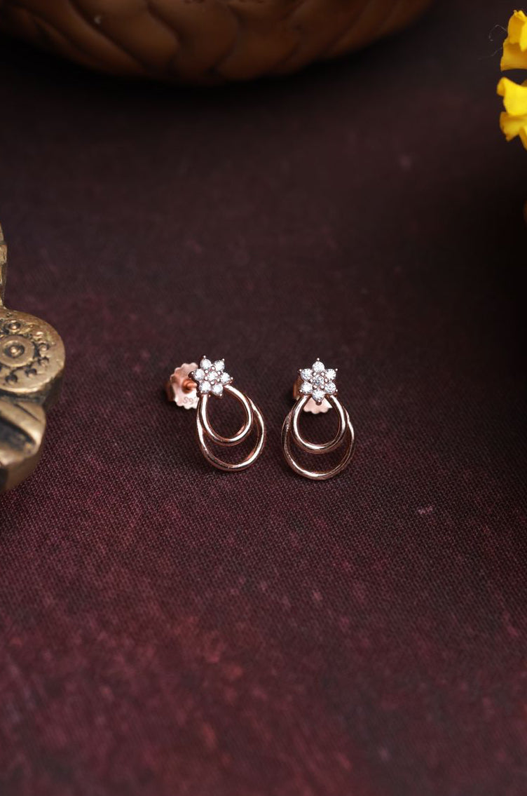 Pair of rose gold earrings on a dark surface with a bowl of round brown sweets and decorative elements.