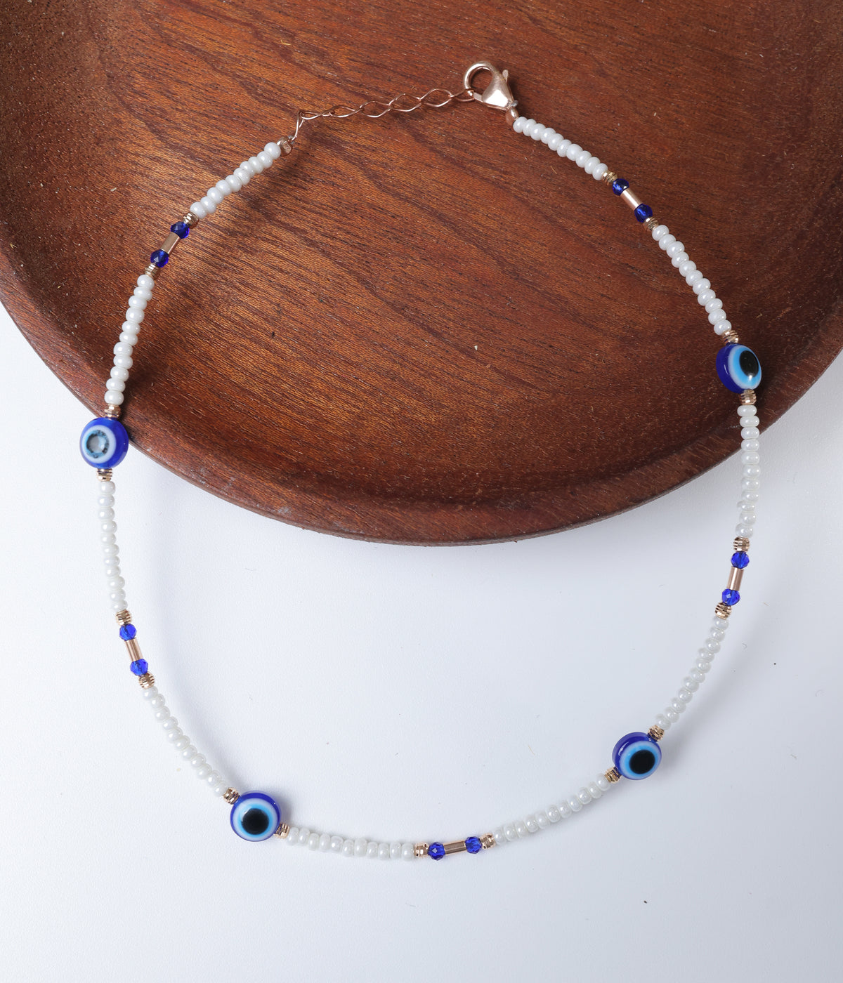 White beaded anklet with blue evil eye charms, cobalt blue crystal accents and rose gold-tone spacers.